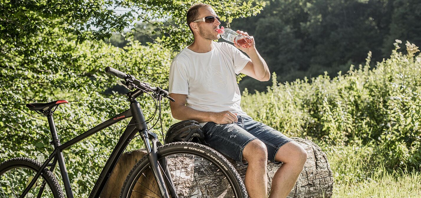 Radfahrer macht Pause im Grünen Radfahrer mit Sonnenbrille trinkt Wasser aus einer Flasche während einer Pause auf einem Baumstamm. Sein schwarzes Mountainbike steht daneben.
