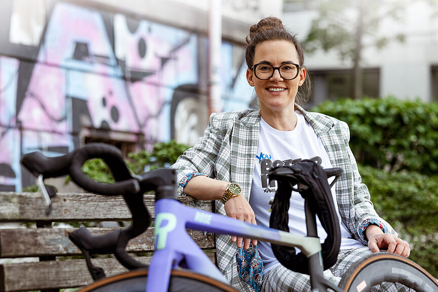 Katja Diehl Eine Frau sitzt entspannt auf einer Holzbank neben einem violetten Rennrad vor einer Graffiti-Wand