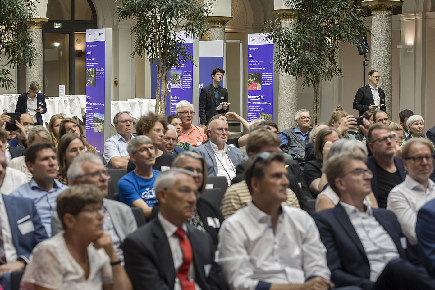 Publikum bei der Veranstaltung Großer Veranstaltungssaal mit vielen Besuchenden in Reihen, überwiegend Männer und Frauen mittleren Alters in Business-Kleidung, im Hintergrund lila Banner zum ADFC-Fahrradklima-Test, Palmen und Säulen in repräsentativem Gebäude, Fotografen dokumentieren die Veranstaltung
